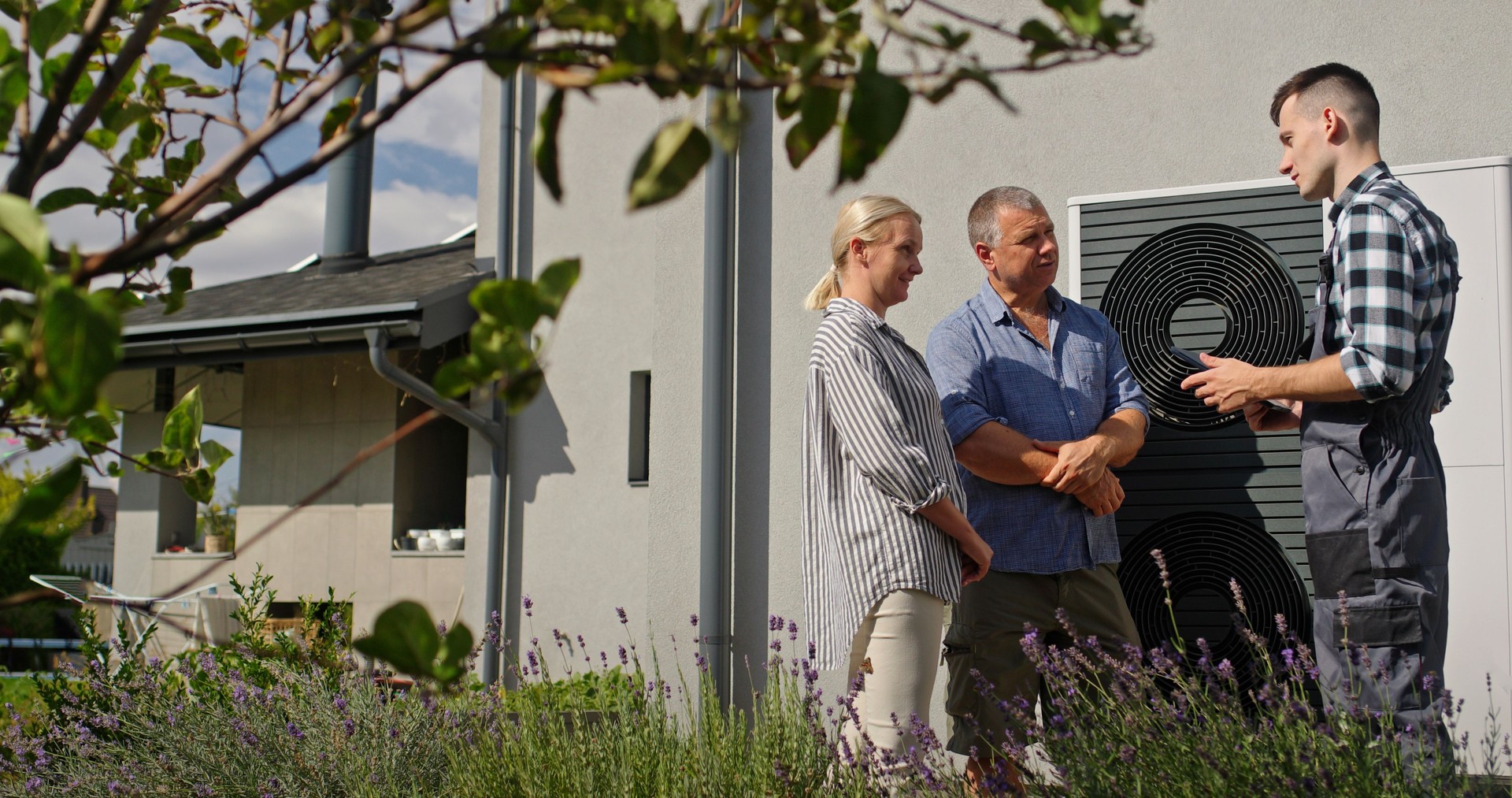 A technician explains heat pump features to a couple using a tablet, standing outdoors near the unit.